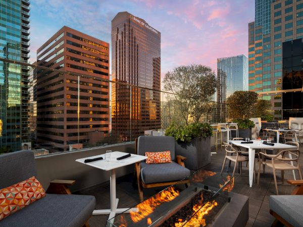A rooftop lounge with modern seating, fire pit, and city skyline backdrop, plush sofas, tables, and greenery at sunset, inviting and chic.