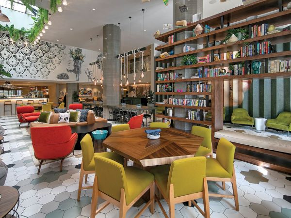 A bright, modern eclectic lounge with hex tiled floor, colorful chairs, hanging plants, a long bookshelf wall, and cozy seating nooks.