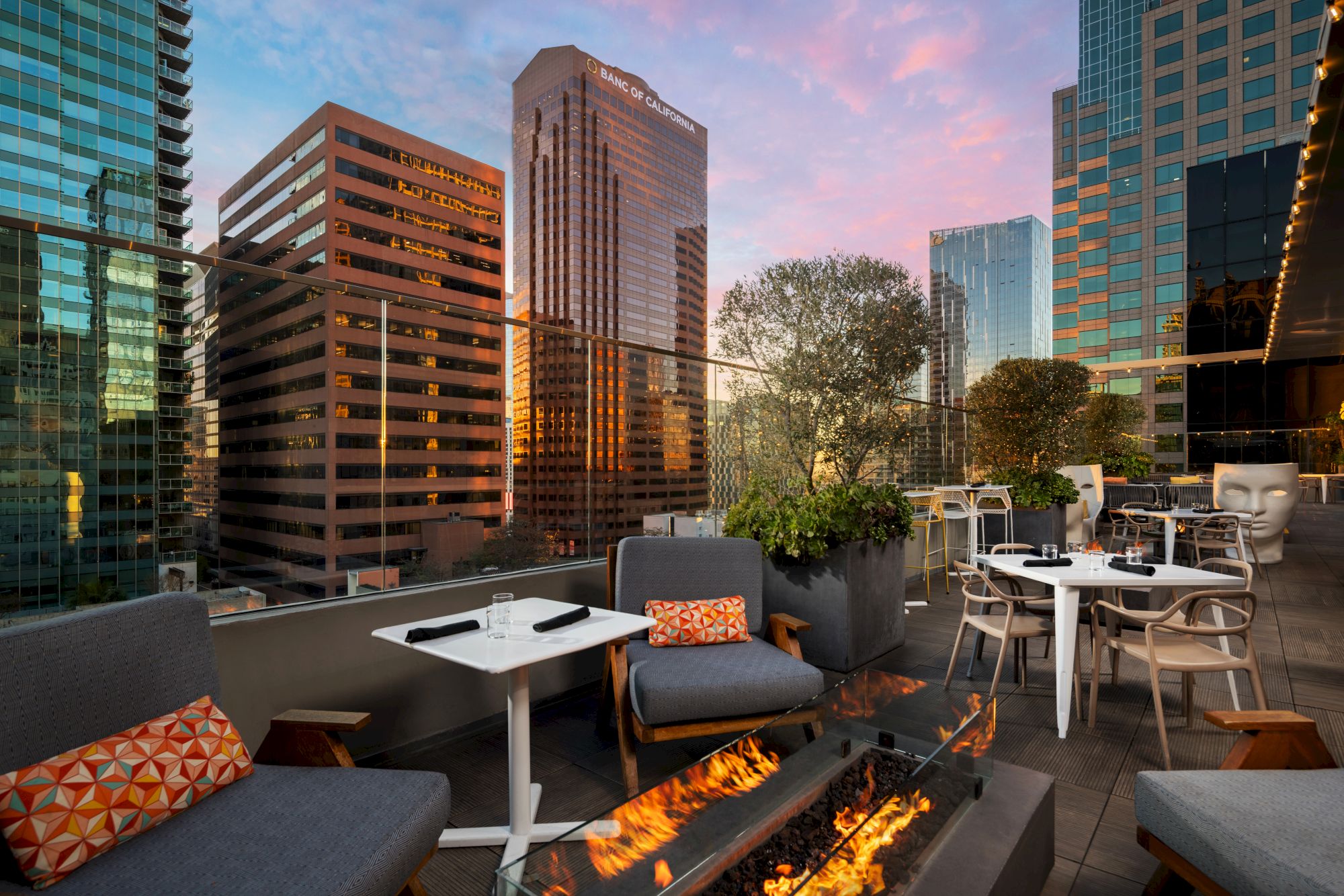 A chic rooftop lounge amid tall glass buildings, cozy couches, fire pits, and small tables with sunset-lit city skyline backdrop.