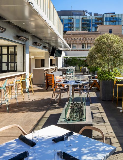 A stylish outdoor cafe terrace with white tables, wooden chairs, bar stools along a counter, potted trees, and modern buildings in the background.