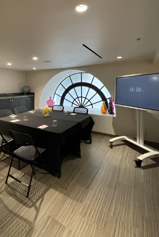A conference room setup with a round window, black-clothed table for six, a large screen on a stand, and colorful plants along the window.