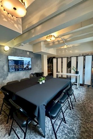 A modern conference room with a long black table, black chairs, overhead lighting, and a wall-mounted screen, plus decorative plants and a partial foldable divider.