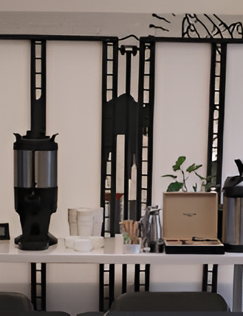 Coffee station setup with a large black coffee urn, cups, a toaster/box server, a water kettle, and a metal rack behind a white table.