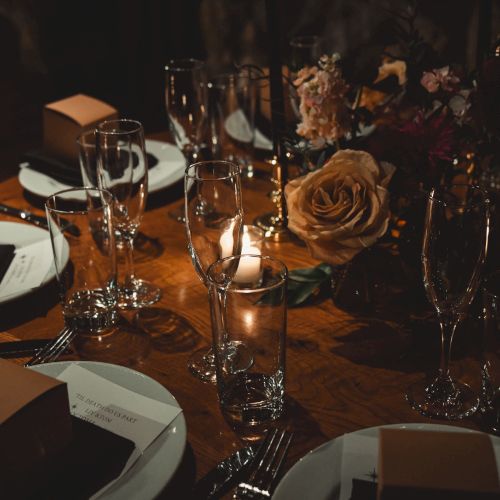 A dimly lit formal dinner table with elegant glassware, white napkins, square brown boxes, candles, and a rose centerpiece, set for guests.