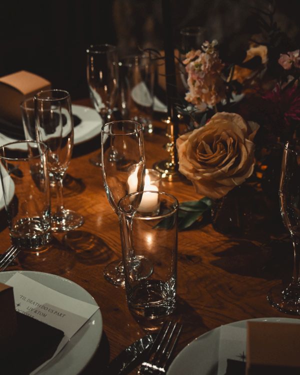 A dimly lit formal dinner table with elegant glassware, white napkins, square brown boxes, candles, and a rose centerpiece, set for guests.