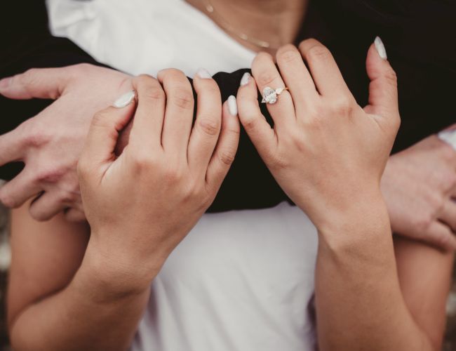 A couple&rsquo;s arms wrapped around each other, hands showing a ring on one finger. The scene feels intimate, romantic, and affectionate.