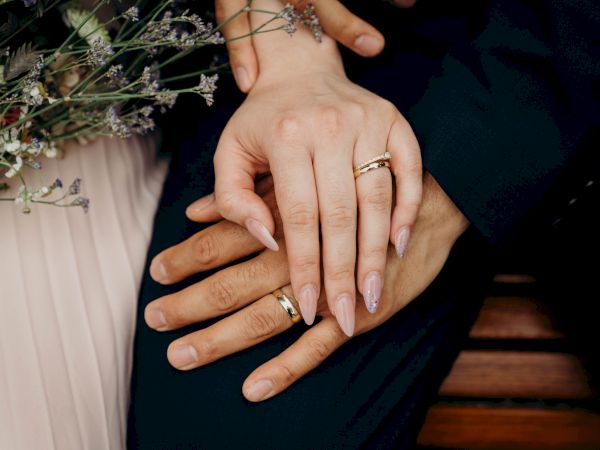 A close-up of two hands with rings resting on a couple&rsquo;s intertwined fingers, wedding bands, delicate nails, and soft floral tones nearby.