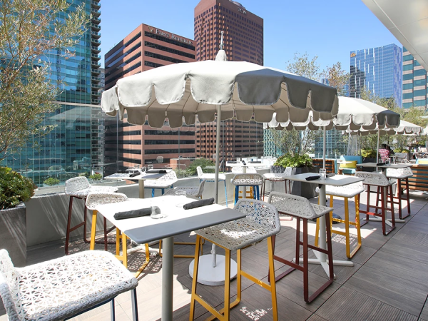 Outdoor city rooftop cafe with white tables, patterned chairs, umbrellas, and high-rise skyline in background.