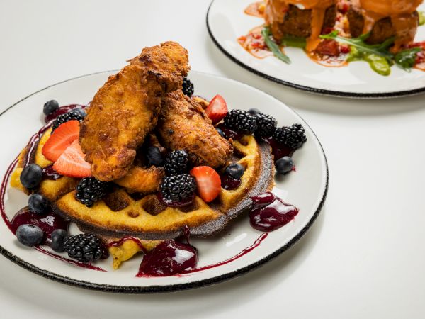 A plate with a fruit-tavored waffle or pancake topped with fried chicken or croquettes, fresh berries, and dollops of berry sauce; another dish in the background. End with a period.