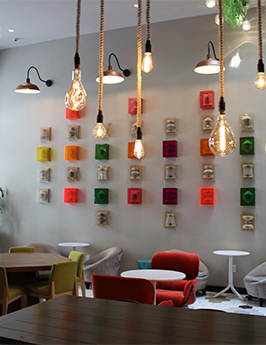A bright, cozy cafe with colorful square blocks on the wall, hanging Edison bulbs, and a mix of red, green, and yellow chairs.