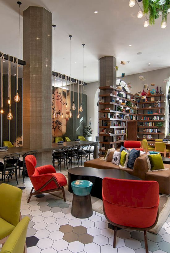 A bright, stylish cafe with colorful chairs, wooden tables, a cozy seating area, hexagon tiled floor, shelves, plants, and large arched windows.