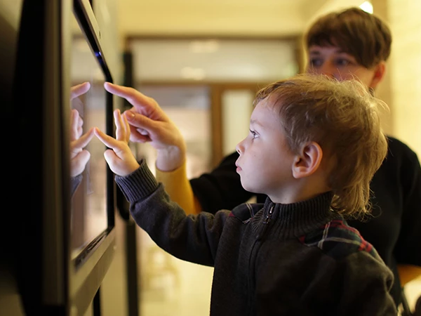 A young boy and an adult look at a touchscreen display, the boy tapping the screen with focused curiosity.