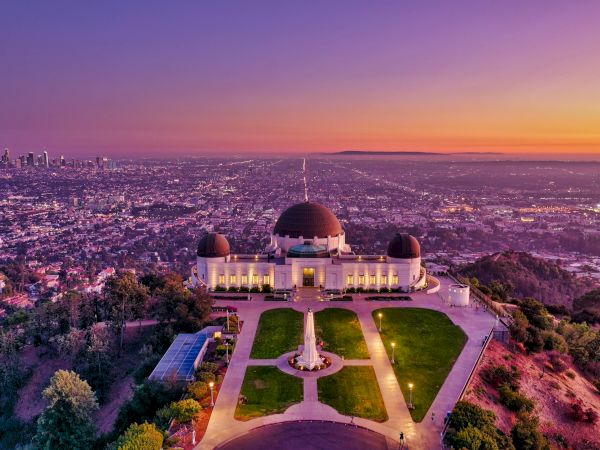 Aerial view of the Griffith Observatory at sunset, with domed towers, symmetrical paths, and a city skyline in the distance, bathed in warm colors.