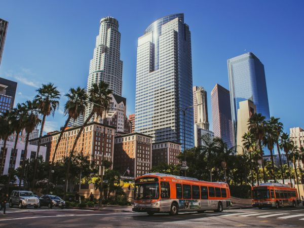 Skyscrapers rise over a sunny downtown with palm trees; a city bus and traffic roam the lively street scene.