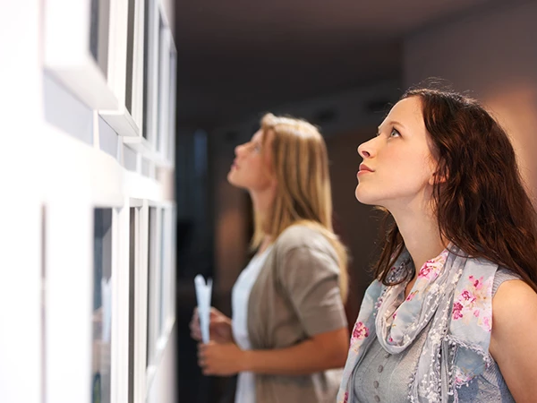 Two women stand indoors, looking intently at framed items on a wall; one in the foreground, the other blurred in the background, both exploring displays.