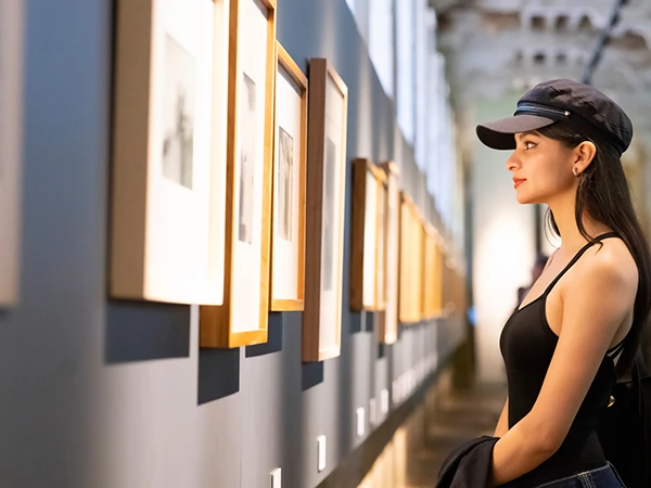 A woman in a black tank top and cap stands in a gallery, admiring framed artworks along a wall.