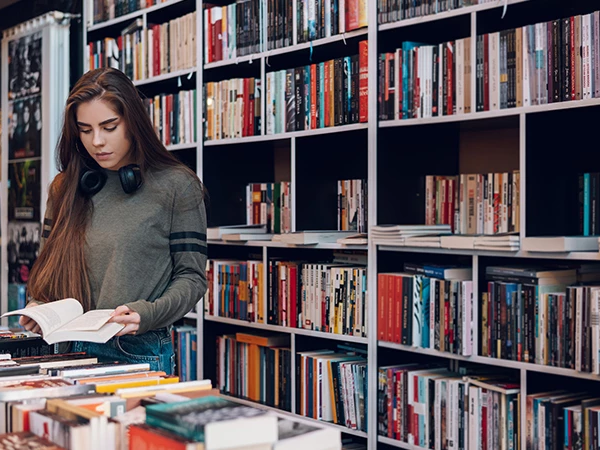 A young woman stands in a bookstore, flipping through a book with shelves of colorful titles surrounding her, listening with headphones.