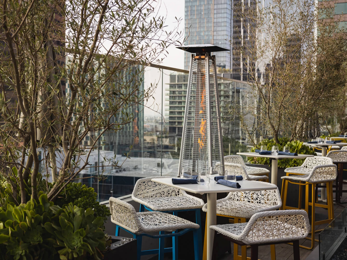 A rooftop restaurant or patio with white patterned chairs around blue tables, tall heaters, planters, and city skyscrapers in the background.