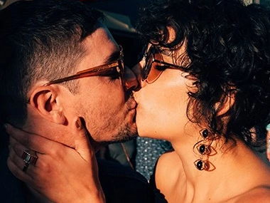 Two people kiss passionately outdoors, close-up on their faces and hands, romantic moment under warm evening light.