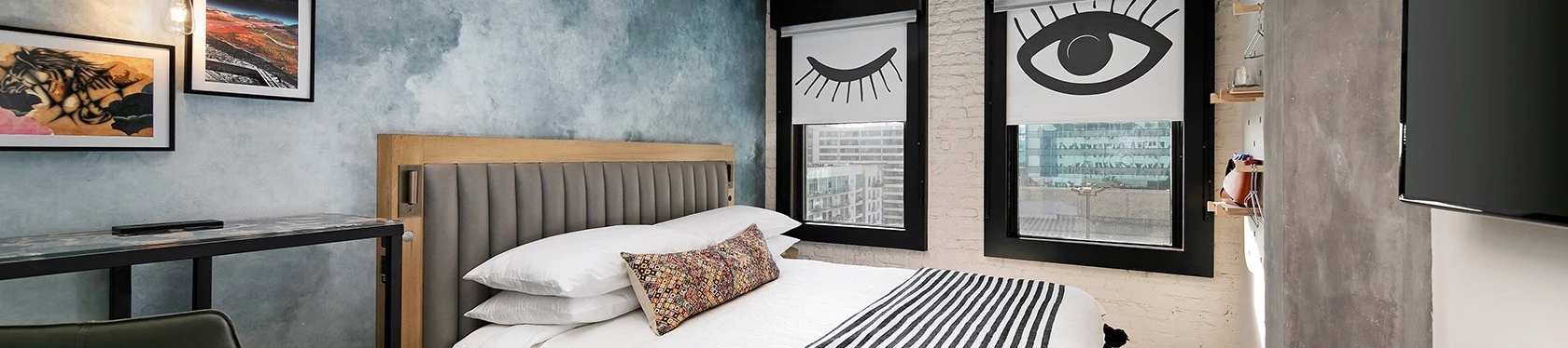 A modern bedroom with a wooden bed, striped bedding, blue textured wall, a desk and chair, two framed eye-themed windows, and a ceiling light fixture, all in a compact, stylish setup.