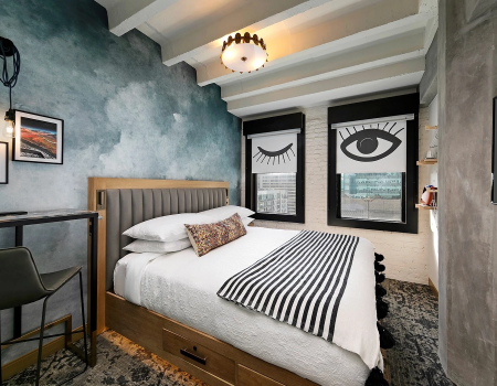 A modern bedroom with a wooden bed, striped bedding, blue textured wall, a desk and chair, two framed eye-themed windows, and a ceiling light fixture, all in a compact, stylish setup.