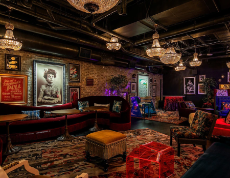 A cozy, eclectic lounge with vintage portraits, plush seating, chandeliers, colorful ottomans, and warm, moody lighting for lounging and socializing.