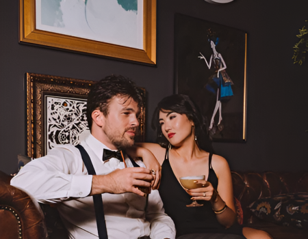 A man in suspenders and a white shirt sits on a leather couch with a woman in a black dress, both holding drinks in a stylish, art-decorated lounge.