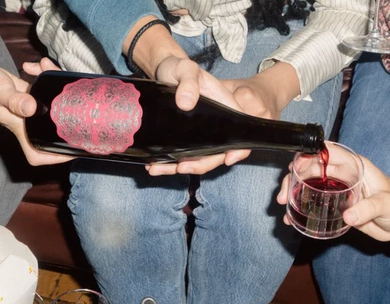 Three people sharing a bottle poured into a shot glass; one person pours, others hold the bottle and glass, casual night out.