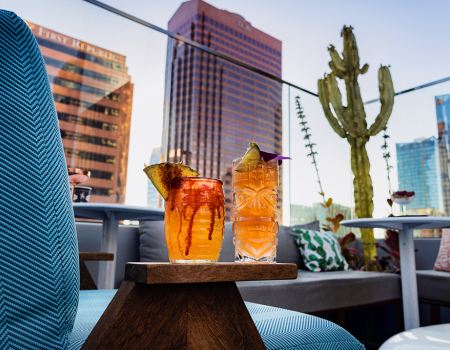 Two tropical cocktails on a wooden tray sit on a rooftop lounge with colorful seating, cactus plants, and city skyscrapers in the background.