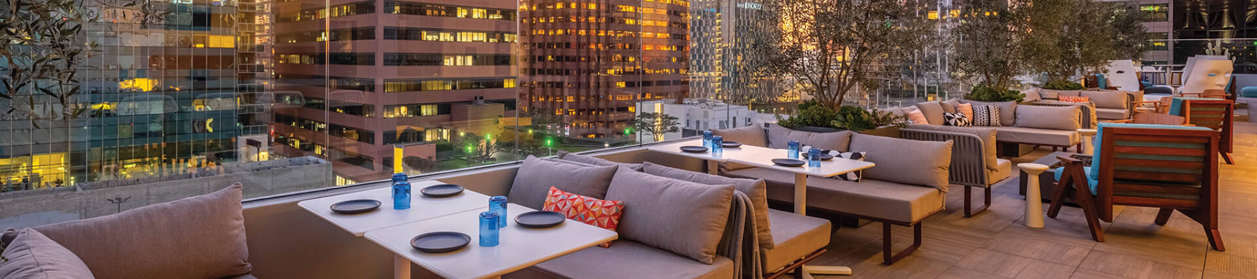 A stylish rooftop dining area with modern couches, small tables set with plates and blue glassware, overlooking a city skyline at dusk.
