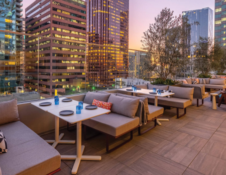 A stylish rooftop dining area with modern couches, small tables set with plates and blue glassware, overlooking a city skyline at dusk.