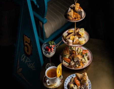 A four-tier tea-time stand piled with pastries, scones, and bites, plus a small cup of tea and a berry dessert on the side, elegant and cozy.