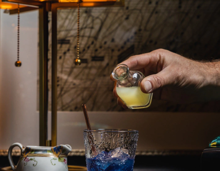A bartender pours a lime wedge into a short glass with blue cocktail, while a hand squeezes a lemon over a drink, in a cozy bar.