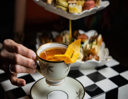 A hand holds a delicate teacup with orange peel garnish, set on a saucer, with a tiered pastries display in the blurred background.
