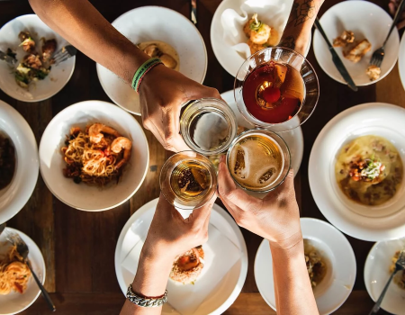 Friends cheers around a table of bowls and plates, clinking drinks in a festive overhead toast ending with a promise of more bites to come.