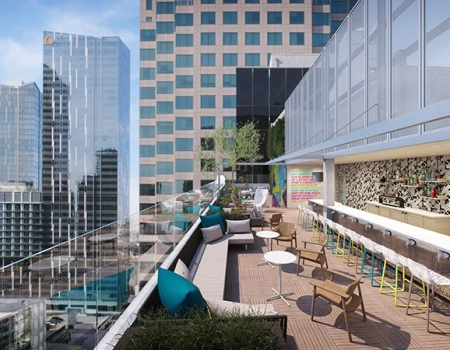 A rooftop terrace with modern chairs and tables, bar area on the right, glass railing, and skyscrapers in the background under a blue sky.