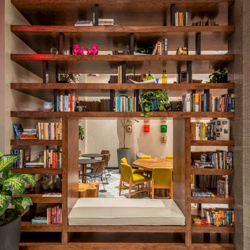 A tall wooden bookshelf frame with a doorway leading to a bright, cozy study area; shelves hold books and plants, hex-tile floor visible.