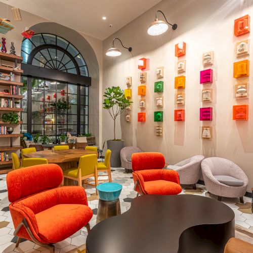 A colorful, cozy library lounge with bright chairs, wall-mounted art blocks, a tall bookshelf, and a large arched window. Top it at 140 characters, ending with a period.