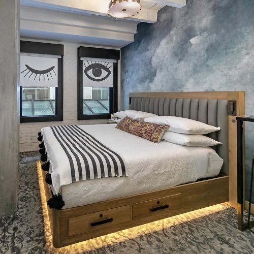 A cozy bedroom with a wooden bed, rustic loft-style frame, warm lighting, a small side table, and framed art on a textured blue wall.