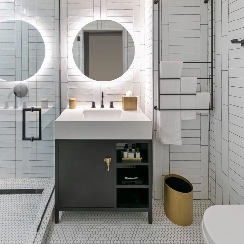 A modern bathroom with a pedestal sink, wall mirror, towel rack, and a washing machine tucked beside a wooden cabinet, all in a light gray and white color scheme.