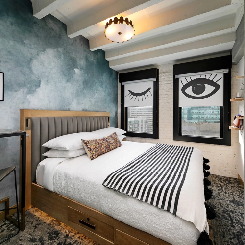 A stylish hotel room with a bed, city view through large windows, chic decor, and a modern workspace; cozy yet contemporary retreat.