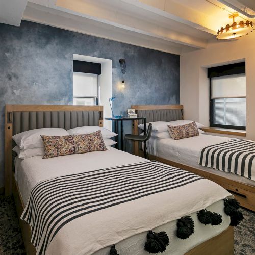 Two double beds in a modern hotel room with a blue accent wall, striped bedding, and a small desk between them.