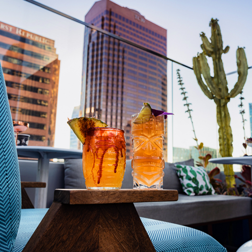 A chic indoor lounge with blue seating, glass railing, and a dramatic skyline view featuring tall towers and a bright blue sky.