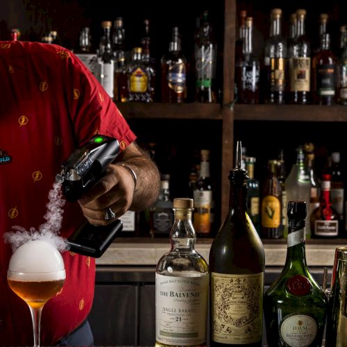 A bartender in a red shirt pours a festive drink for a customer against a well-lit bar backdrop, with bottles lining the shelves, sparkling lights.