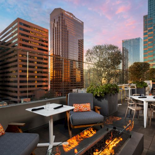 A stylish rooftop lounge with modern seating, a glowing fire pit, and city skyline views at sunset, perfect for cocktails and conversation.