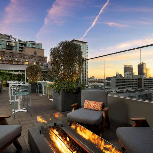A stylish rooftop lounge with comfy seating, a modern fire pit, potted trees, and a city skyline at sunset.