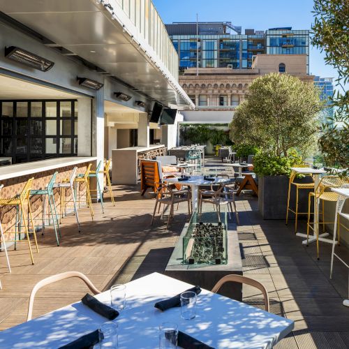 A sunlit rooftop restaurant with tables, chairs, and a bar overlooking the city, perfect for outdoor dining.