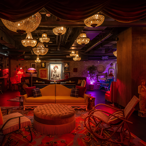 A cozy, dimly lit lounge with red seating, warm lamps, and glowing torches; a small stage or bar area in the background.