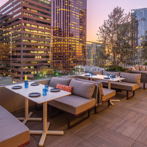 A stylish rooftop lounge at dusk with warm string lights, modern seating, and a skyline view of high-rise buildings.