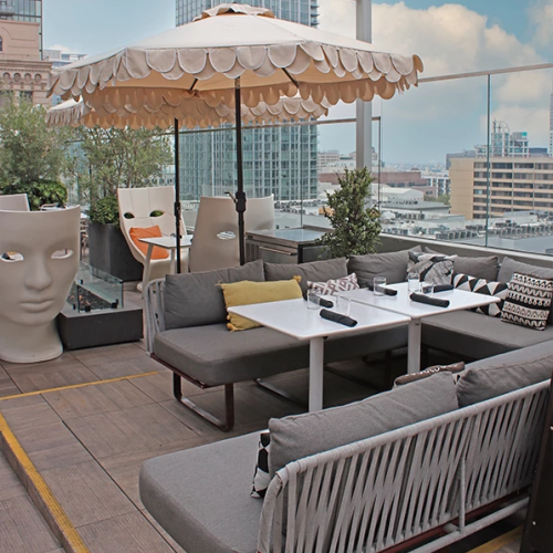 A rooftop city scene with a striped umbrella over a seating area, chain-link fence, scaffolding, and distant buildings.
